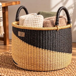May include: A round woven basket featuring a two-tone design with black and natural tan fibres. The basket has two black wrapped handles and a small rectangular leather-like patch on the front. It is filled with several folded textured towels.