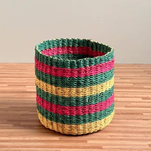 Può includere: Un cesto cilindrico intrecciato caratterizzato da strisce orizzontali in colori alternati verde scuro, rosa acceso e giallo oro. Il cesto presenta una superficie intrecciata e testurizzata con un'apertura superiore e un motivo uniforme esterno.