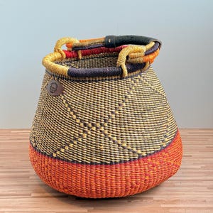 May include: A woven basket featuring a wide, rounded base in vibrant orange, transitioning to a dark brown and yellow patterned body. It includes a thick, black leather-wrapped handle and a small, circular brown leather patch with a logo.