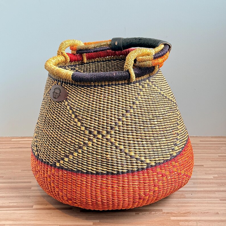 May include: A woven basket featuring a wide, rounded base in vibrant orange, transitioning to a dark brown and yellow patterned body. It includes a thick, black leather-wrapped handle and a small, circular brown leather patch with a logo.