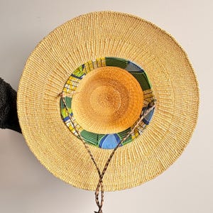 Puede incluir: Un sombrero de paja de ala ancha con una textura tejida en color beige natural. El interior de la copa presenta un forro de tela colorido con un patr&oacute;n geom&eacute;trico en tonos verdes, azules y amarillos. Un cord&oacute;n trenzado marr&oacute;n est&aacute; unido al ala.