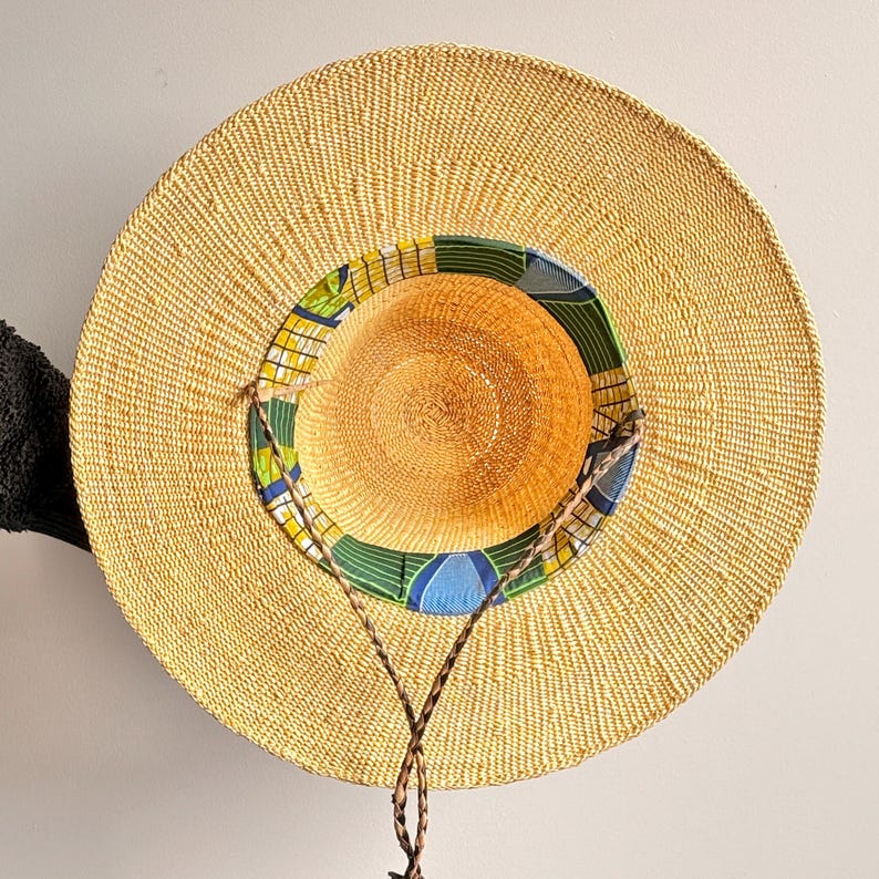 Puede incluir: Un sombrero de paja de ala ancha con una textura tejida en color beige natural. El interior de la copa presenta un forro de tela colorido con un patr&oacute;n geom&eacute;trico en tonos verdes, azules y amarillos. Un cord&oacute;n trenzado marr&oacute;n est&aacute; unido al ala.