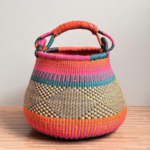 Könnte beinhalten: Ein runder, handgeflochtener Korb mit einem lebendigen, mehrfarbigen Muster in Rosa, Orange, Blaugrün und natürlichen Strohtönen. Der Korb hat eine weite Öffnung und einen einzelnen, stabilen, umwickelten Griff in passenden Orange- und Rosatönen.