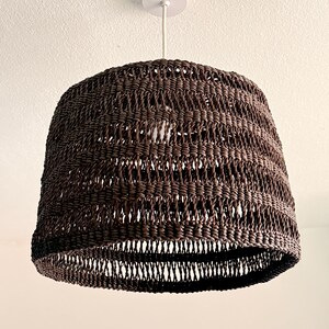 K&ouml;nnte beinhalten: Eine gewebte, braune Pendelleuchte. Der Lampenschirm hat eine Glockenform und ein offenes Webmuster. Sie h&auml;ngt an einem wei&szlig;en Kabel und einer Deckenhalterung. Die Leuchte ist von der Decke abgeh&auml;ngt.