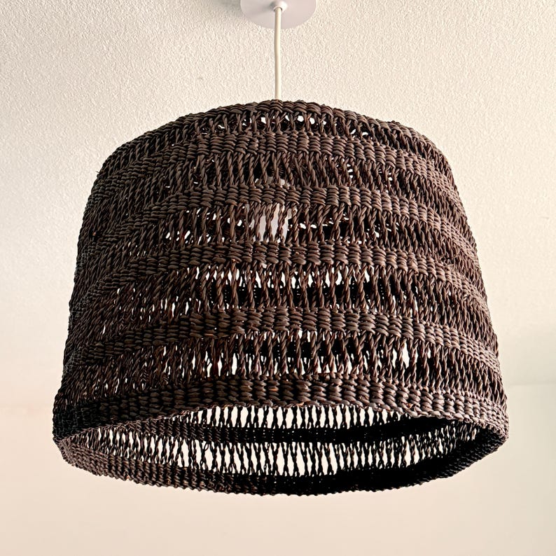 K&ouml;nnte beinhalten: Eine gewebte, braune Pendelleuchte. Der Lampenschirm hat eine Glockenform und ein offenes Webmuster. Sie h&auml;ngt an einem wei&szlig;en Kabel und einer Deckenhalterung. Die Leuchte ist von der Decke abgeh&auml;ngt.