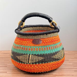 May include: A round woven basket featuring horizontal bands of bright orange, yellow, teal, and black. The basket has a thick black rim and a sturdy black handle wrapped in light-colored cord. A small, oval brown leather patch is centered on the front.