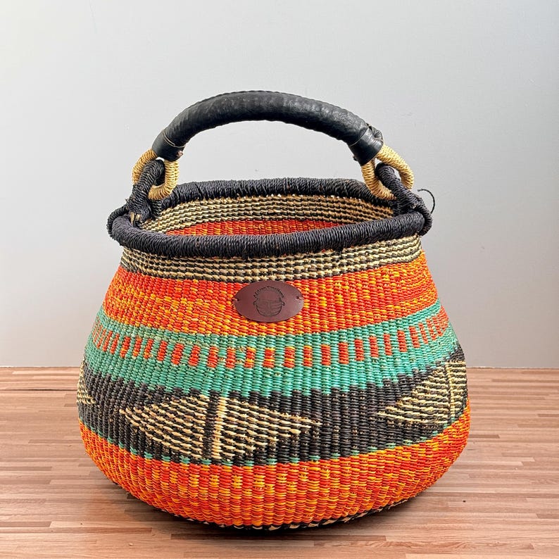 May include: A round woven basket featuring horizontal bands of bright orange, yellow, teal, and black. The basket has a thick black rim and a sturdy black handle wrapped in light-colored cord. A small, oval brown leather patch is centered on the front.