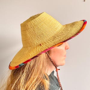 Puede incluir: Un sombrero de paja de ala ancha con una textura tejida en color amarillo natural. La parte inferior del ala presenta un forro de tela vibrante y multicolor con patrones en rojo, naranja y azul. Una correa trenzada cuelga del lateral.