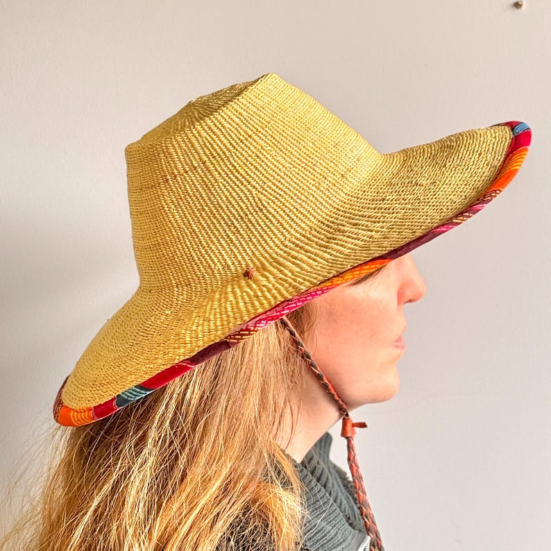 Puede incluir: Un sombrero de paja de ala ancha con una textura tejida en color amarillo natural. La parte inferior del ala presenta un forro de tela vibrante y multicolor con patrones en rojo, naranja y azul. Una correa trenzada cuelga del lateral.