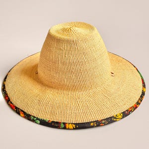 Puede incluir: Un sombrero de paja de ala ancha con una copa alta y redondeada, de textura beige finamente tejida. El borde exterior del ala est&aacute; rematado con un ribete de tela negra que presenta un vibrante estampado floral multicolor en tonos rojos, amarillos y verdes.