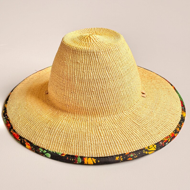 Puede incluir: Un sombrero de paja de ala ancha con una copa alta y redondeada, de textura beige finamente tejida. El borde exterior del ala est&aacute; rematado con un ribete de tela negra que presenta un vibrante estampado floral multicolor en tonos rojos, amarillos y verdes.