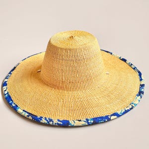 Puede incluir: Un sombrero de paja de ala ancha con una textura tejida en color tostado natural y una copa plana. El borde exterior del ala est&aacute; rematado con un ribete de tela estampado en azul y blanco con un dise&ntilde;o floral o org&aacute;nico abstracto.