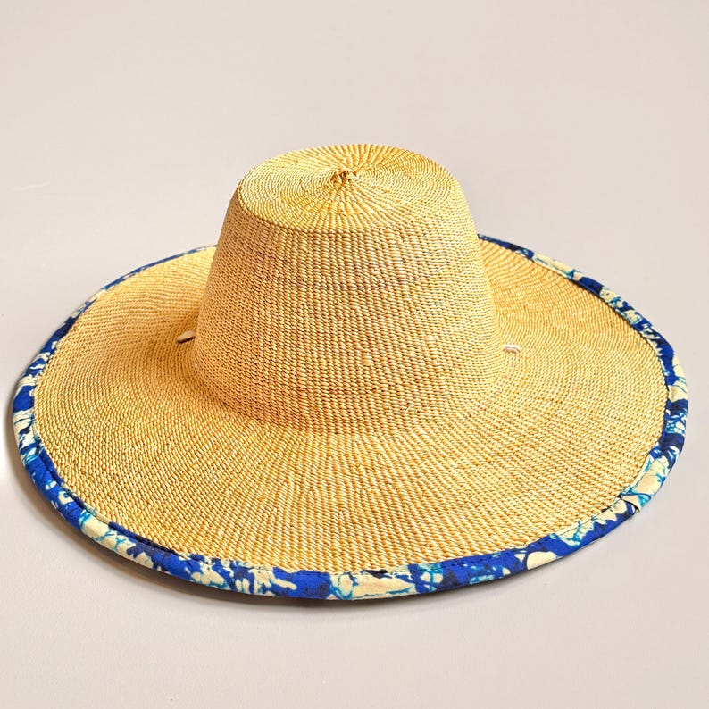 Puede incluir: Un sombrero de paja de ala ancha con una textura tejida en color tostado natural y una copa plana. El borde exterior del ala est&aacute; rematado con un ribete de tela estampado en azul y blanco con un dise&ntilde;o floral o org&aacute;nico abstracto.