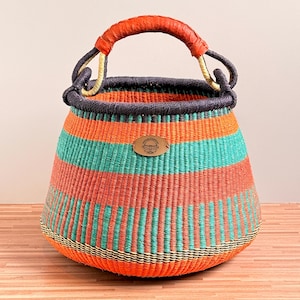 Op de afbeelding: Een handgeweven mand met horizontale strepen in feloranje en turquoise. De mand heeft een donkerblauwe rand en een omwikkeld oranje lederen handvat. Een ovaal metalen label met een gegraveerd logo bevindt zich in het midden aan de voorzijde.