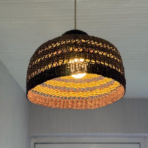 K&ouml;nnte beinhalten: Gewebte Pendelleuchte mit braun-schwarzem Muster. Die Leuchte hat eine runde Form und ein gewebtes Design. Die Gl&uuml;hbirne ist sichtbar und strahlt ein warmes Licht aus.