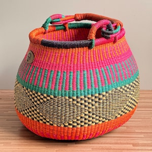 May include: A round, hand-woven basket featuring a vibrant pattern of horizontal stripes in bright pink, orange, teal, and black. The basket has two woven handles at the top, one wrapped in brown leather, and a small circular brown logo tag on the side.