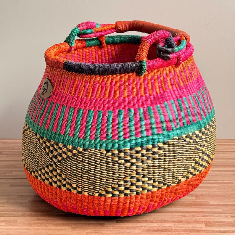 May include: A round, hand-woven basket featuring a vibrant pattern of horizontal stripes in bright pink, orange, teal, and black. The basket has two woven handles at the top, one wrapped in brown leather, and a small circular brown logo tag on the side.