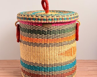 May include: A cylindrical woven basket with a lid featuring a red handle and side loops. The basket displays horizontal bands of yellow, green, orange, teal, and brown. A small oval metal plaque is centred on the upper portion of the basket body.