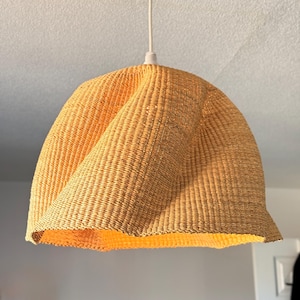 K&ouml;nnte beinhalten: Ein gewebter, hellgelber Lampenschirm h&auml;ngt an einem wei&szlig;en Kabel. Der Lampenschirm hat ein einzigartiges, verdrehtes Design, und die Textur des gewebten Materials ist sichtbar. Das Licht der Gl&uuml;hbirne im Inneren beleuchtet die Unterseite des Schirms.