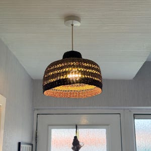 K&ouml;nnte beinhalten: Eine gewebte Pendelleuchte mit braunem und schwarzem Lampenschirm h&auml;ngt von einer wei&szlig;en Decke. Das Licht ist eingeschaltet und beleuchtet das Innere des Schirms. Die Leuchte wird von einem d&uuml;nnen, cremefarbenen Kabel abgeh&auml;ngt.