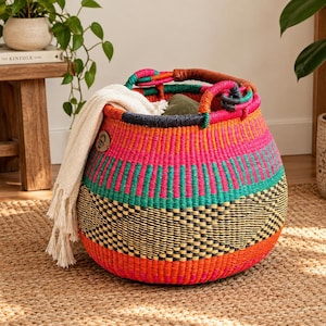 May include: A round, hand-woven basket featuring vibrant horizontal stripes in hot pink, teal, and orange, with a black geometric pattern near the base. It has two leather-wrapped handles and contains a cream-colored, fringed textile throw.