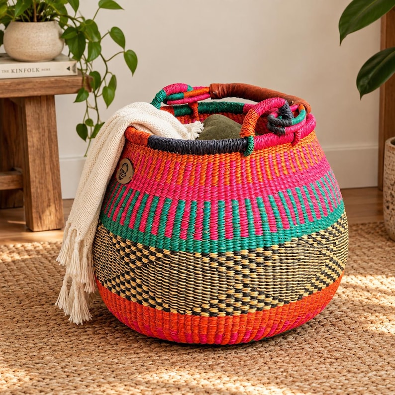 May include: A round, hand-woven basket featuring vibrant horizontal stripes in hot pink, teal, and orange, with a black geometric pattern near the base. It has two leather-wrapped handles and contains a cream-colored, fringed textile throw.