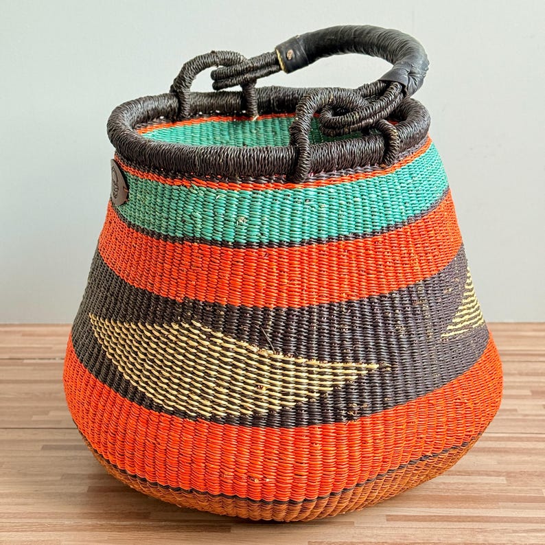 May include: A round, hand-woven basket featuring horizontal stripes in vibrant orange, dark brown, and teal. The basket has a wide, open top with a dark brown woven rim and a single, sturdy, leather-wrapped handle attached to the top edge.
