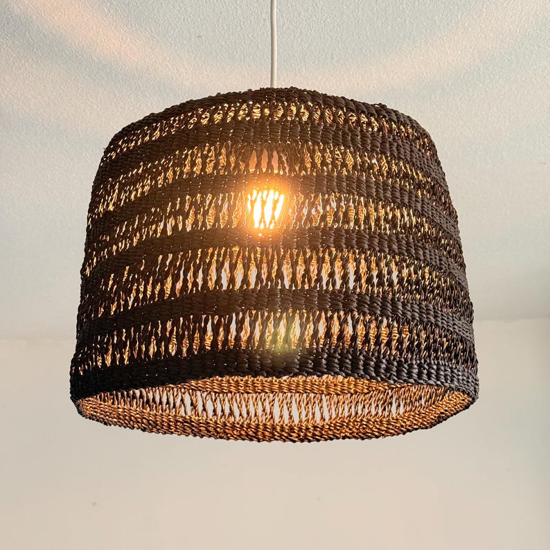 K&ouml;nnte beinhalten: Eine schwarze, gewebte Pendelleuchte mit konischer Form. Die Leuchte h&auml;ngt an einem wei&szlig;en Kabel und strahlt ein warmes Licht aus. Das gewebte Design l&auml;sst das Licht durchscheinen und erzeugt einen strukturierten Effekt.
