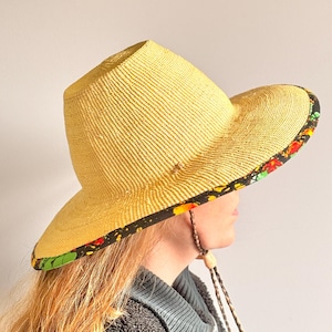 Puede incluir: Un sombrero de paja amarillo con copa plana y ala ancha. La parte inferior del ala presenta un borde de tela negra decorado con un vibrante estampado floral en rojo, naranja y verde. Una correa trenzada de color marr&oacute;n est&aacute; sujeta al sombrero.