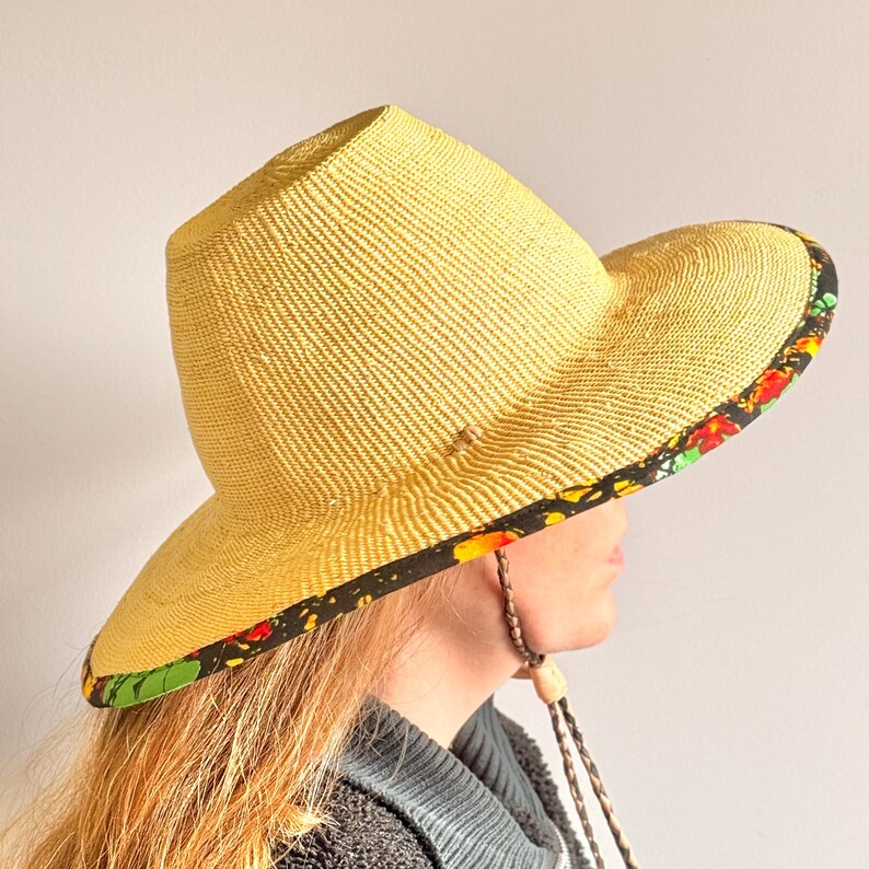 Puede incluir: Un sombrero de paja amarillo con copa plana y ala ancha. La parte inferior del ala presenta un borde de tela negra decorado con un vibrante estampado floral en rojo, naranja y verde. Una correa trenzada de color marr&oacute;n est&aacute; sujeta al sombrero.