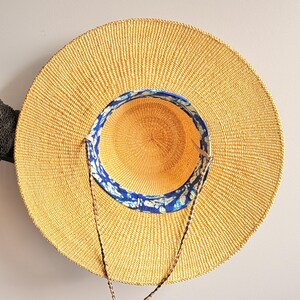 Puede incluir: Un sombrero de paja de ala ancha con una textura tejida en color beige natural. El interior de la copa presenta un forro de tela con un patr&oacute;n en azul y blanco. Una fina correa de barbilla de cord&oacute;n trenzado marr&oacute;n se sujeta a los lados.