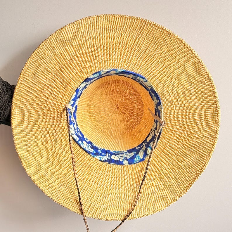 Puede incluir: Un sombrero de paja de ala ancha con una textura tejida en color beige natural. El interior de la copa presenta un forro de tela con un patr&oacute;n en azul y blanco. Una fina correa de barbilla de cord&oacute;n trenzado marr&oacute;n se sujeta a los lados.