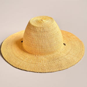 Puede incluir: Un sombrero de paja de ala ancha en color amarillo claro con una copa redondeada y una superficie tejida texturizada. El sombrero presenta dos peque&ntilde;os detalles en marr&oacute;n oscuro en los lados opuestos donde el ala se une a la copa.