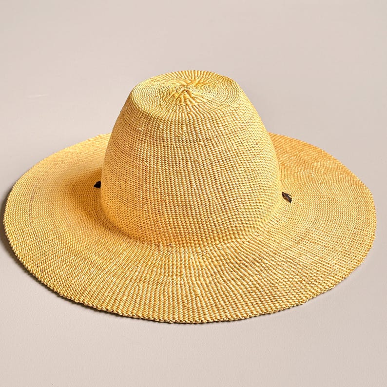 Puede incluir: Un sombrero de paja de ala ancha en color amarillo claro con una copa redondeada y una superficie tejida texturizada. El sombrero presenta dos peque&ntilde;os detalles en marr&oacute;n oscuro en los lados opuestos donde el ala se une a la copa.