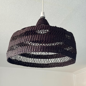 K&ouml;nnte beinhalten: Eine dunkelbraune gewebte Pendelleuchte. Der Lampenschirm hat eine abgerundete, glockenf&ouml;rmige Form mit einem gewebten Muster. Das Licht h&auml;ngt an einem wei&szlig;en Kabel und einer wei&szlig;en Deckenleuchte. Die Leuchte ist ein dekoratives Element f&uuml;r die Inneneinrichtung.