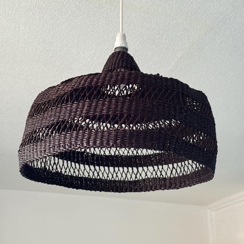 K&ouml;nnte beinhalten: Eine dunkelbraune gewebte Pendelleuchte. Der Lampenschirm hat eine abgerundete, glockenf&ouml;rmige Form mit einem gewebten Muster. Das Licht h&auml;ngt an einem wei&szlig;en Kabel und einer wei&szlig;en Deckenleuchte. Die Leuchte ist ein dekoratives Element f&uuml;r die Inneneinrichtung.