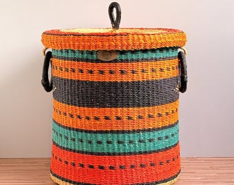 May include: A cylindrical woven storage basket with a lid, featuring horizontal stripes in orange, black, and teal. The basket includes two black side handles and a black handle on the lid, with a small oval leather-like brand tag on the front.