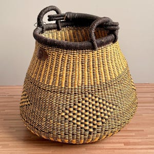 May include: A round, hand-woven basket featuring a geometric pattern in alternating black and natural yellow straw. The basket has a thick, dark brown wrapped rim and two sturdy, matching dark brown handles attached to the top edge.