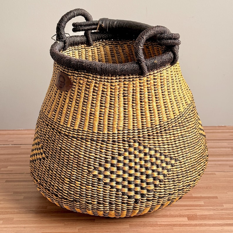 May include: A round, hand-woven basket featuring a geometric pattern in alternating black and natural yellow straw. The basket has a thick, dark brown wrapped rim and two sturdy, matching dark brown handles attached to the top edge.
