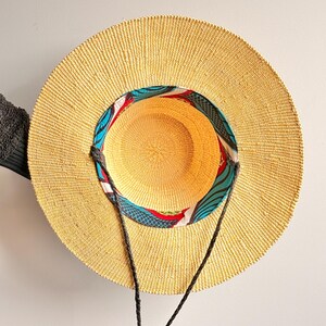 Puede incluir: Un sombrero de paja de ala ancha con una textura tejida en color tostado natural. El interior de la copa presenta un forro de tela colorido con un patr&oacute;n abstracto en azul, rojo y blanco. Dos cordones trenzados de color marr&oacute;n oscuro est&aacute;n unidos.
