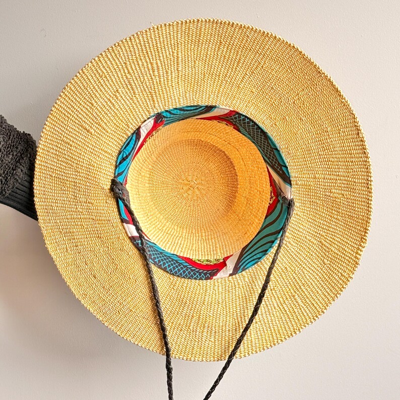 Puede incluir: Un sombrero de paja de ala ancha con una textura tejida en color tostado natural. El interior de la copa presenta un forro de tela colorido con un patr&oacute;n abstracto en azul, rojo y blanco. Dos cordones trenzados de color marr&oacute;n oscuro est&aacute;n unidos.