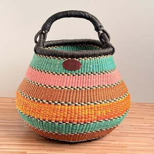 May include: A round woven basket featuring horizontal stripes in teal, pink, orange, and yellow. The basket has a dark brown wrapped handle and a dark brown rim. A small, oval-shaped brown leather patch is centred on the upper portion of the basket.
