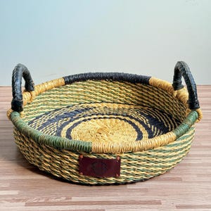 May include: A round, shallow woven basket featuring a coiled pattern in shades of yellow, green, and dark blue. The basket includes two sturdy black handles attached to the sides and a rectangular brown leather patch centred on the front exterior.