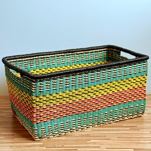 Pode incluir: Um cesto de armazenamento retangular com um padrão entrançado de riscas horizontais vibrantes em amarelo, laranja e turquesa, rematado com um rebordo preto sólido e pegas integradas no topo, feito de material sintético durável.