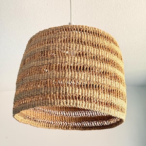 K&ouml;nnte beinhalten: Eine gewebte Pendelleuchte in hellem Braun. Der Lampenschirm hat eine bauchige Form und ist an einem wei&szlig;en Kabel aufgeh&auml;ngt. Das gewebte Design l&auml;sst das Licht durchscheinen und erzeugt eine warme Atmosph&auml;re.