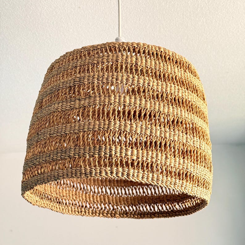 K&ouml;nnte beinhalten: Eine gewebte Pendelleuchte in hellem Braun. Der Lampenschirm hat eine bauchige Form und ist an einem wei&szlig;en Kabel aufgeh&auml;ngt. Das gewebte Design l&auml;sst das Licht durchscheinen und erzeugt eine warme Atmosph&auml;re.