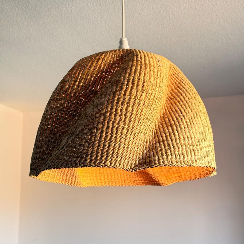 K&ouml;nnte beinhalten: Eine gewebte Pendelleuchte mit einem einzigartigen, geschwungenen Design. Der Lampenschirm besteht aus einem hellbraunen, strukturierten Material und h&auml;ngt an einem wei&szlig;en Kabel. Das Innere des Schirms strahlt ein warmes, einladendes Licht aus.