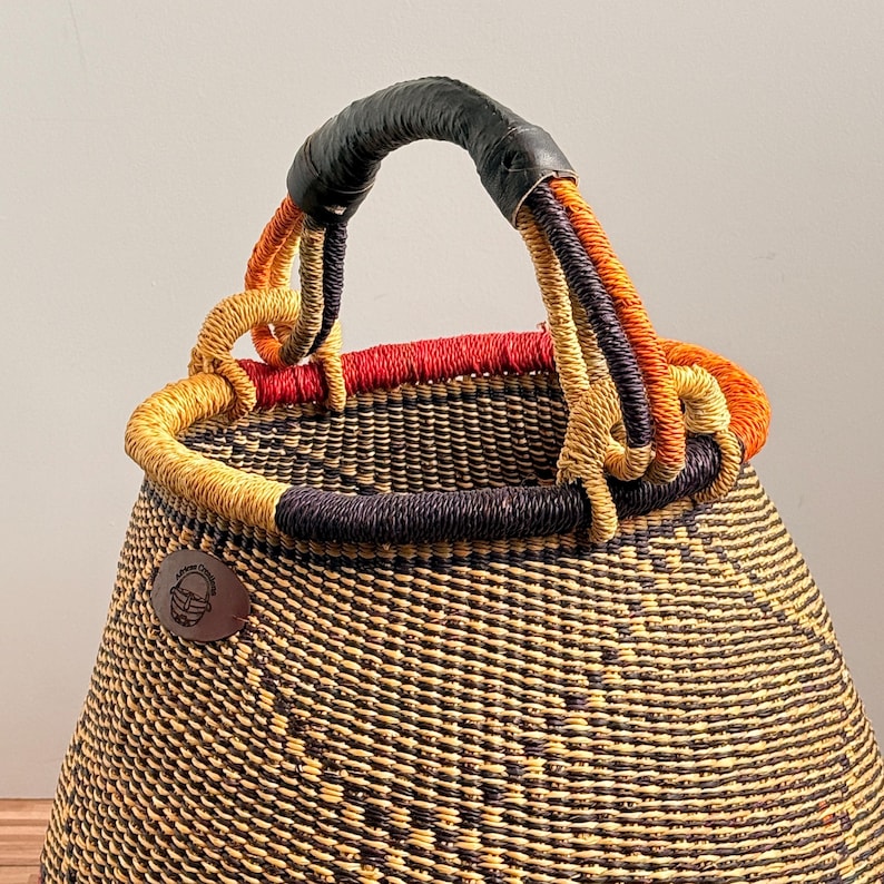 May include: A woven basket with a conical shape, featuring a dark brown and yellow textured pattern. The rim is accented with red, orange, and purple fibers. It has a black leather-wrapped handle and a small brown circular logo patch on the side.