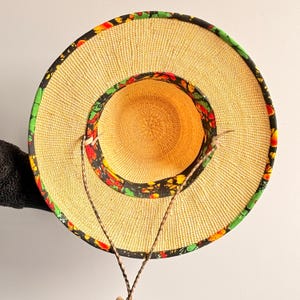 Puede incluir: Un sombrero de paja de ala ancha con una textura tejida en tono bronceado natural, acentuado con un borde de tela negra decorado con un vibrante patr&oacute;n floral en rojo, amarillo y verde. Incluye una fina correa trenzada marr&oacute;n.