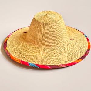 Puede incluir: Un sombrero de paja de ala ancha con una copa alta y redondeada de textura beige tejida. El borde exterior del ala est&aacute; rematado con un ribete de tela vibrante que presenta rayas diagonales en tonos rojo brillante, naranja, morado y azul cerceta.